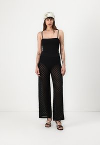 Crna uska bodysuit uparen s prozirnim, uzorkastim hlačama širokih nogavica. Model nosi bijelu maramu s uzorkom i crne sandale s remenčićima.