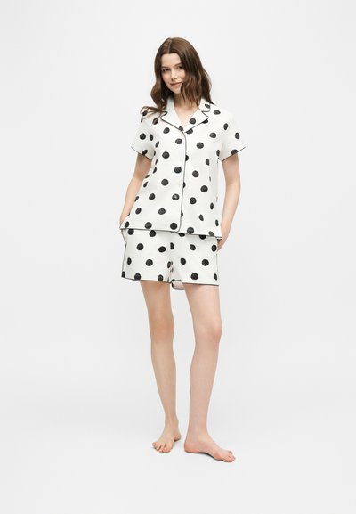Femme debout pieds nus portant un haut blanc à manches courtes boutonné et un short assorti à gros pois noirs sur un fond uni.