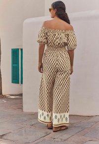 Off-shoulder, mönstrad jumpsuit i beige och brun, med en smockad midja och vida ben med dekorativt trim och svarta lövmotiv.