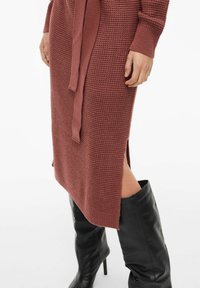 Strikket kjole i rustfarge med teksturert mønster, med belte som knytes i livet, sidesplitt og lange ermer. Brukes sammen med svarte knelange støvler.