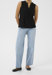 Blouse noire sans manches avec motif de découpes en forme de diamants, associée à un jean large bleu clair et des chaussures plates noires.