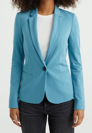 Person iført en tætsiddende lyseblå blazer over en hvid skjorte og mørke jeans, står med armene afslappet ned langs siden.