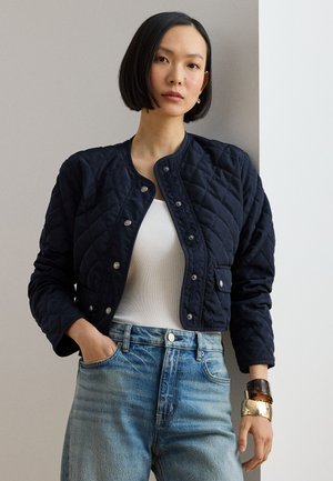 Kvinde med lige sort bob-frisure iført en quiltet marineblå jakke, hvid top, blå jeans og store armbånd på hendes højre håndled.