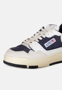 Baskets blanches, marine et beige avec une tige en suede, des panneaux texturés et des lacets plats, dotées d'une patch logo et d'un emblème sur le côté.
