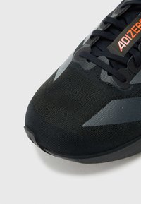 Zapato deportivo negro con parte superior de tela texturizada. Presenta detalles reflectantes y una puntera moldeada. Los cordones son oscuros, con un logo "ADIZERO".