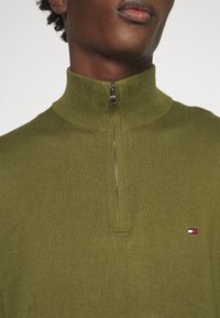 Gros plan d'une personne portant un pull en maille côtelée vert olive avec un col demi-fermeture éclair et un petit logo de drapeau rouge, blanc et bleu sur la poitrine.