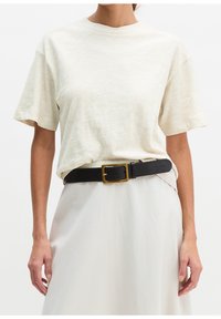 Femme portant un t-shirt uni beige clair à manches courtes, rentré dans une jupe blanche avec une ceinture noire avec une boucle carrée dorée.