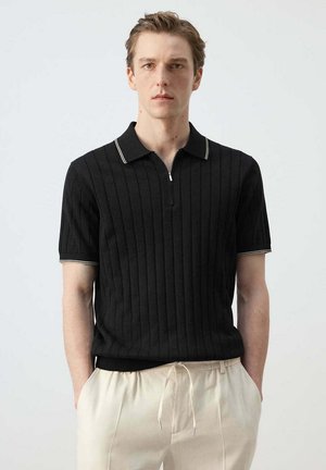 Hombre que lleva una camiseta tipo polo de manga corta negra con ribete blanco y pantalones beige claro con cordón, de pie con las manos en los bolsillos.