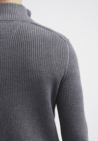 Primo piano di una persona che indossa un maglione grigio lavorato a maglia con una texture a coste, cucitura della spalla visibile e colletto alto.