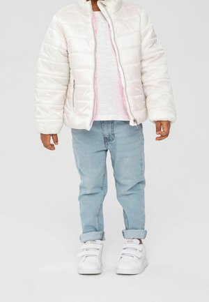 Lapsi, jolla on valkoinen toppatakki vaaleanpunaisen paidan päällä, vaaleansiniset käännetyt farkut ja valkoiset Adidas-tennarit, joissa on tarranauhat.