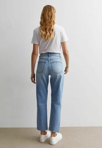 Mujer con cabello rubio ondulado que lleva una camiseta blanca, jeans de tiro alto en un azul claro y zapatillas blancas, de pie frente a un fondo liso.