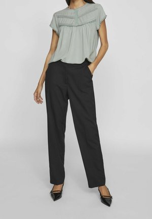 Femme portant un chemisier à manches courtes gris clair avec des plis et un pantalon noir droit, posant avec une main dans la poche.