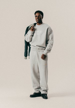 Mann in hellgrauem Sweatshirt und Jogginghose, der eine dunkle Jacke über eine Schulter hält, vor einem schlichten hellen Hintergrund.