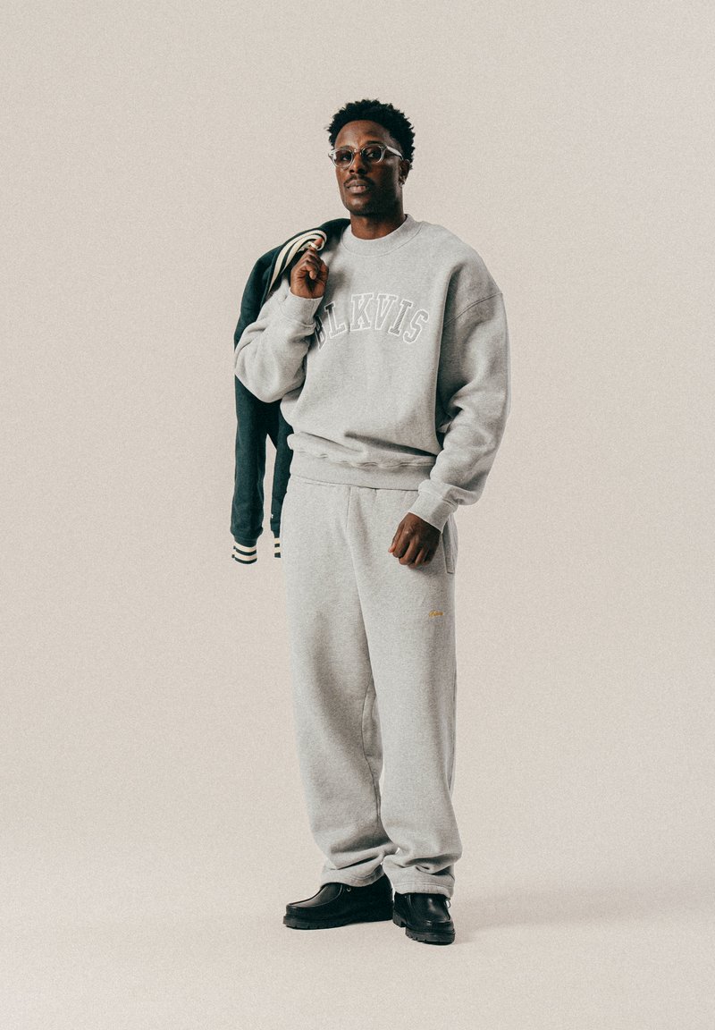 Mężczyzna w jasnoszarej bluzie dresowej i dresach, trzymający ciemną kurtkę na jednym ramieniu, na jasnym jednolitym tle.