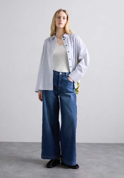 Gestreiftes Hemd über einem weißen Tanktop, kombiniert mit hochgeschnittenen, weit geschnittenen blauen Jeans und schwarzen Schuhen. Gelbe Tasche sichtbar.