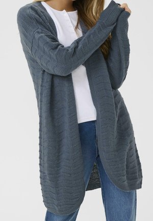 Femme portant un long cardigan gris tricoté par-dessus une chemise blanche boutonnée et un jean bleu, ajustant une manche avec les deux mains.