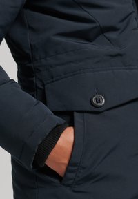 Marine blauwe pufferjack met een textuurstof, voorzien van een zijzak, zwarte knoopsluiting en ribgebreide manchetdetails.