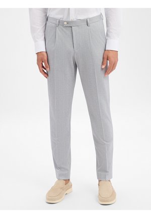 Uomo che indossa pantaloni eleganti grigi chiaro a righe sottili, camicia bianca con bottoni e scarpe beige senza lacci, su sfondo bianco semplice.