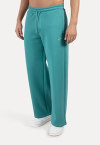 Turkusowe spodnie dresowe ze ściągaczem w talii, bocznymi kieszeniami oraz kontrastowym logo marki na udzie, w połączeniu z białymi trampkami.