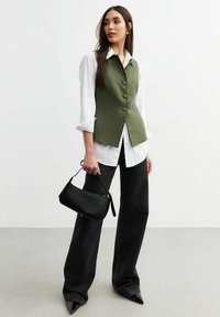 Chaleco verde con botones sobre una camisa blanca, combinado con jeans de pierna ancha negros y tacones negros puntiagudos. Sosteniendo un pequeño bolso negro.