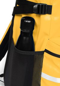 Mochila amarilla con un bolsillo lateral de malla negra y una botella de agua negra cilíndrica. Acento reflectante en la mochila.