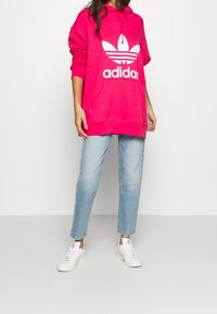 Osoba v jasně růžové mikině Adidas, světlých modrých džínách a bílých teniskách stojí na jednobarevném světlém pozadí.