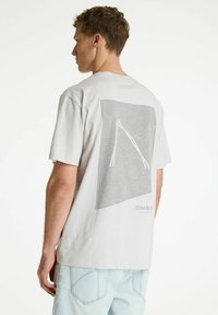 Joven mirando hacia la izquierda, vistiendo una camiseta gris claro con un diseño topográfico abstracto blanco y jeans azul claro.