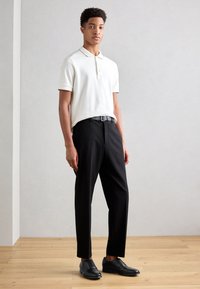 Camisa polo blanca de manga corta con cuello, combinada con pantalones negros y zapatos negros con cordones, sobre un suelo de madera con un fondo claro.