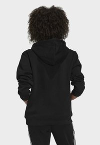 Svart hoodie som visas bakifrån, med en dragsko i huvan, mjuk textur och ribbad manschett. I kombination med svarta träningsbyxor med vita ränder.