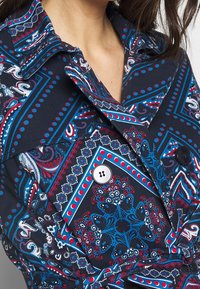Koszula w wzory z granatową bazą, ozdobiona czerwonymi i turkusowymi wzorami paisley oraz geometrycznymi. Ciekawym elementem są duży kołnierz i guziki.