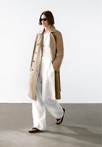 Beżowy płaszcz trencz noszony na białej bluzce i luźnych białych spodniach, dopełniony czarnymi klapkami i okularami przeciwsłonecznymi, na minimalistycznym tle.