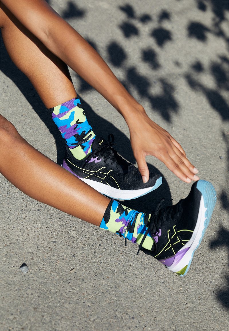 Persona estirando las piernas sobre el pavimento, vistiendo calcetines coloridos y zapatillas de correr negras con acentos morados y azules.
