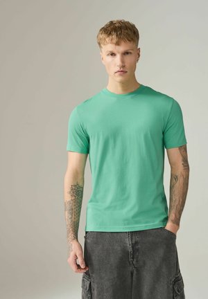 Tânăr cu păr blond scurt, purtând un tricou verde și pantaloni cargo negri, arătând tatuajele de pe ambele antebrațe, stând cu o mână în buzunar.