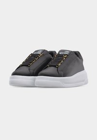 Μαύρα sneakers Moschino με χρυσά μεταλλικά στοιχεία, λευκές χοντρές σόλες και λεπτομέρειες με το λογότυπο στη γλώσσα και στις πλευρές, παρουσιασμένα σε ανοιχτό γκρι φόντο.