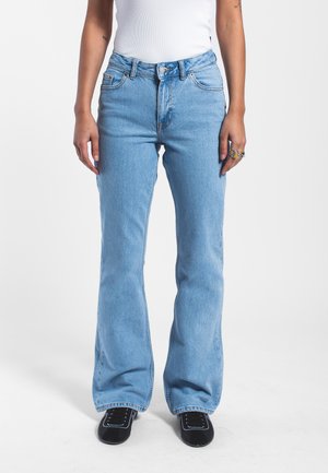 Personne portant un jean flare taille haute bleu clair, un haut blanc, des chaussures noires et des bagues à la main droite sur un fond uni.