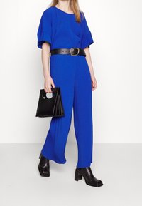 Kobieta w królewskim niebieskim prążkowanym kombinezonie z czarnym paskiem, czarnych botkach na obcasach i trzymająca czarną torebkę z okrągłymi uchwytami.