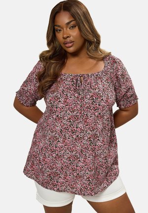 Nainen, jolla on pitkät laineikkaat hiukset ja joka pukeutuu vaaleanpunaiseen kukalliseen lyhythihaiseen paitaan sekä valkoisiin shortseihin, seisoo kädet selän takana.