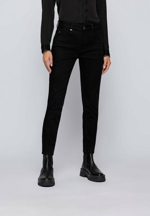 Frau trägt schwarze Slim-Fit-Jeans, schwarzen langärmligen Oberteil mit Spitzenbesatz und schwarze Leder-Stiefeletten, steht vor einem schlichten Hintergrund.