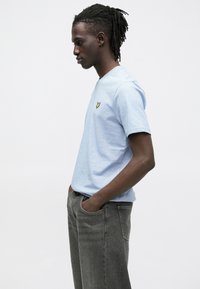 Světle modré bavlněné tričko s kulatým výstřihem, krátkými rukávy a malým žlutým logem na hrudi, kombinované se šedými džínami.