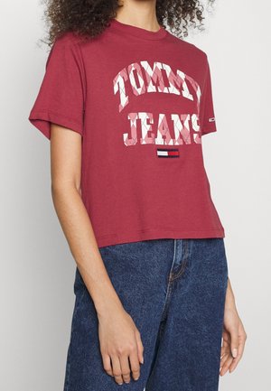 Kobieta ubrana w krótką czerwoną koszulkę Tommy Jeans z białym kratkowanym napisem oraz niebieskie dżinsy z wysokim stanem.