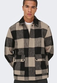 Rutig ulljacka i beige och svart med krage, framknäppning med tryckknappar, två påsydda fickor och en texturerad yta. Fyrkantigt mönster över hela.