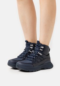 Marineblauwe wandelschoenen met een gevoerde kraag, een gestructureerde zool, metalen onderdelen en veters met contrasterende stiksels.