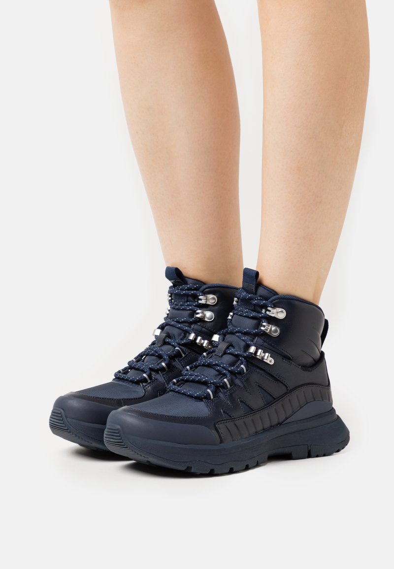 Marineblauwe wandelschoenen met een gevoerde kraag, een gestructureerde zool, metalen onderdelen en veters met contrasterende stiksels.