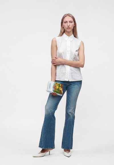 Femme portant un chemisier blanc sans manches et un jean bleu tient un livre intitulé Toscana, debout devant un fond blanc uni.