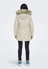 Personne portant un parka beige avec une capuche en fourrure, un jean noir et des baskets bleu et blanc, se tenant de dos sur un fond uni.