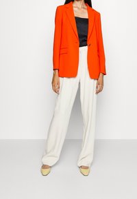 Oranžový blazer se strukturovanými rameny a zapínáním na jeden knoflík, spárovaný s béžovými kalhotami s vysokým pasem a světle žlutými botami na blokovém podpatku.