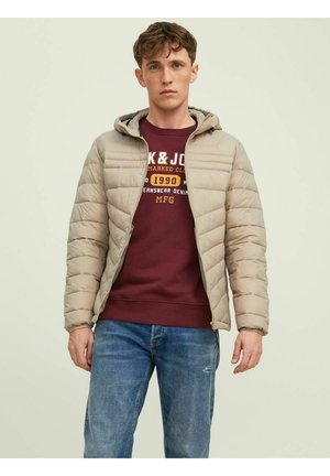 Beige boblejakke med hette, horisontal quiltedesign og glidelås foran. Båret over en maroon genser og blå jeans. Løs passform.