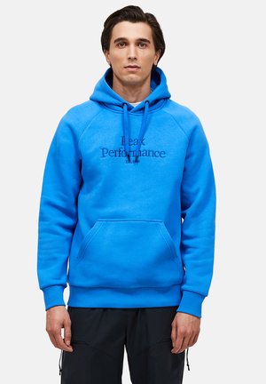Homme portant un sweat à capuche bleu vif avec le texte "Peak Performance Est. 1986", debout devant un fond clair uni, les mains détendues le long du corps.