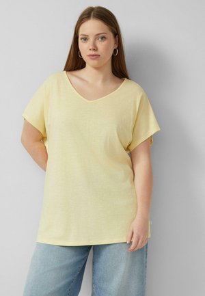 Młoda kobieta z prostymi długimi włosami nosi bladożółtą luźną bluzkę z dekoltem w szpic oraz jasnoniebieskie dżinsy, stoi na jednolitym szarym tle.