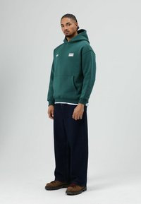 Groen capuchontrui met een voorzak van het kangoeroetype, gecombineerd met donkerblauwe wijde jeans en bruine laarzen met rode accenten.